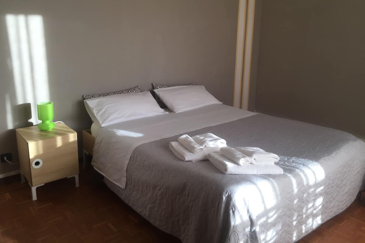 A neatly made bed with white linens and a grey quilt, a bedside table with a green lamp, and a wooden floor.
