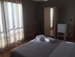 A hotel room with a bed, a wooden wardrobe, a radiator, a chair, and a small white cabinet. - Casa Salice (Racconigi)