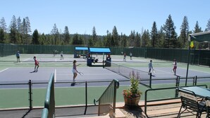 Un campo da tennis all'aperto con diversi giocatori, un'area ombreggiata per sedersi e una recinzione intorno.