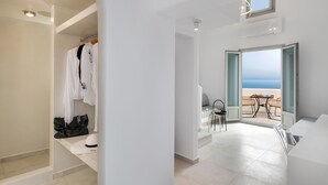 A modern hotel room with a balcony overlooking the sea, a seating area with a table and chairs, and a walk-in closet with hanging clothes and a suitcase.