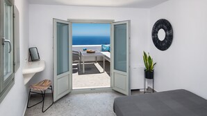 A bedroom with a bed, a small table with a chair, a potted plant, and a view of the sea through an open door.