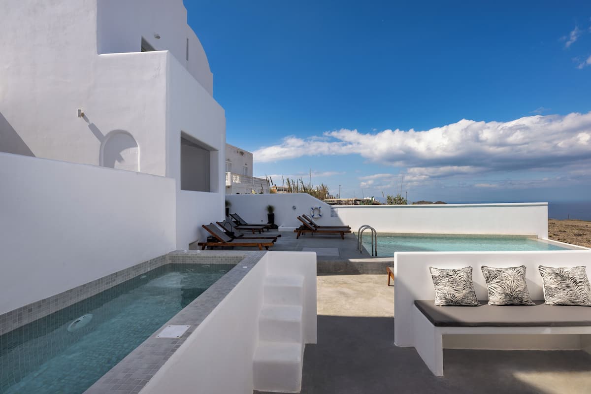Un área de piscina moderna al aire libre con un edificio blanco, sillas de descanso y una zona de estar con cojines.
