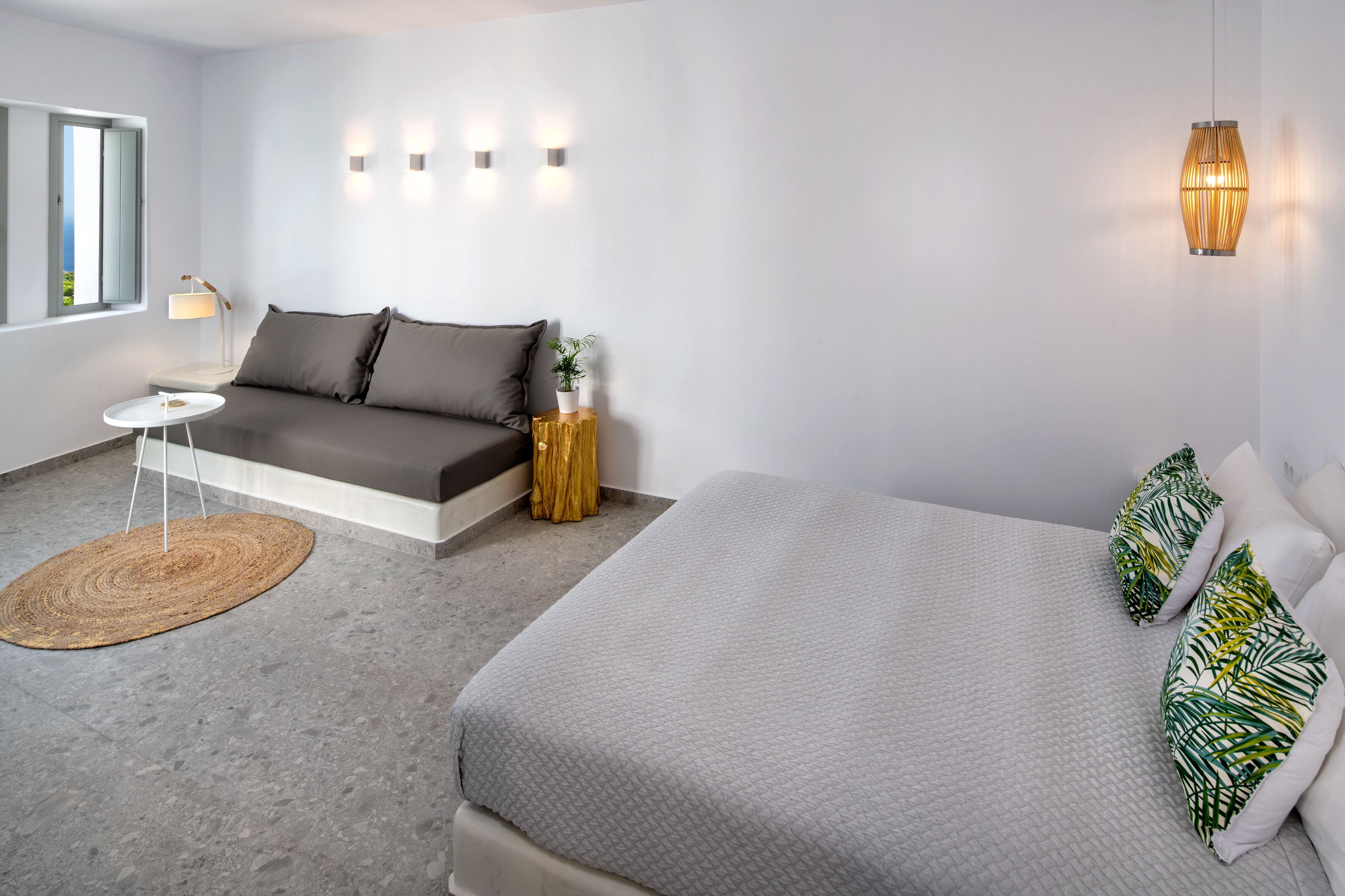 A modern bedroom with a grey sofa, a bed with green and white patterned pillows, a small round table, and a wall-mounted lamp.
