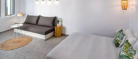 A modern bedroom with a grey sofa, a bed with green and white patterned pillows, a small round table, and a wall-mounted lamp.