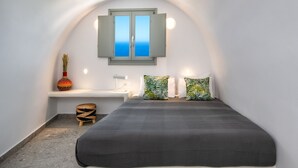 A bedroom with a bed, a desk, a chair, a small stool, a vase, and a window with shutters.