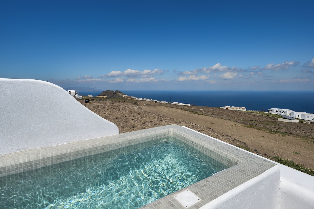 Vista clara de una piscina con borde de azulejos, una playa arenosa y una lejana vista al mar y edificios blancos.