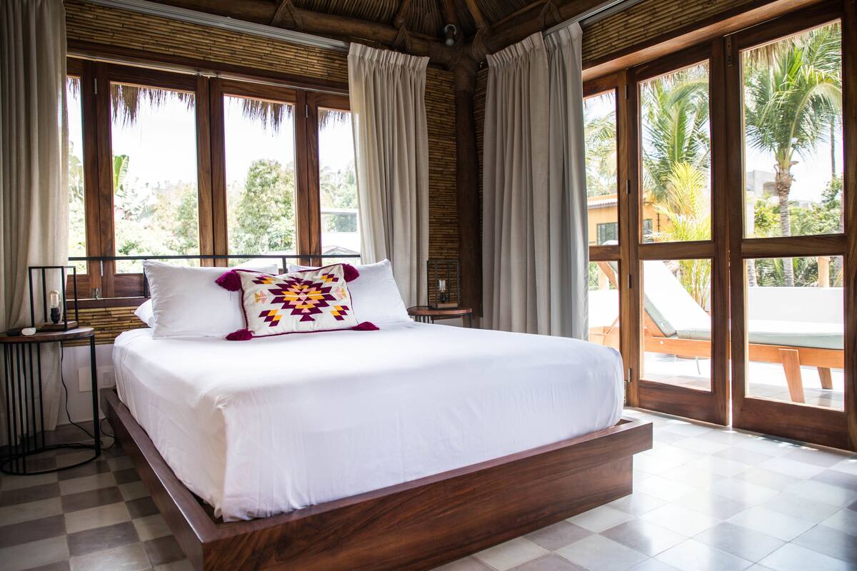 A large bed with white linens and a colorful pillow, a bedside table, and a view of palm trees through large windows.
