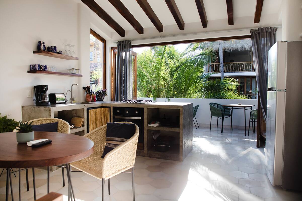 A modern kitchen with a dining area, a balcony with a view of palm trees, and a small outdoor seating area.