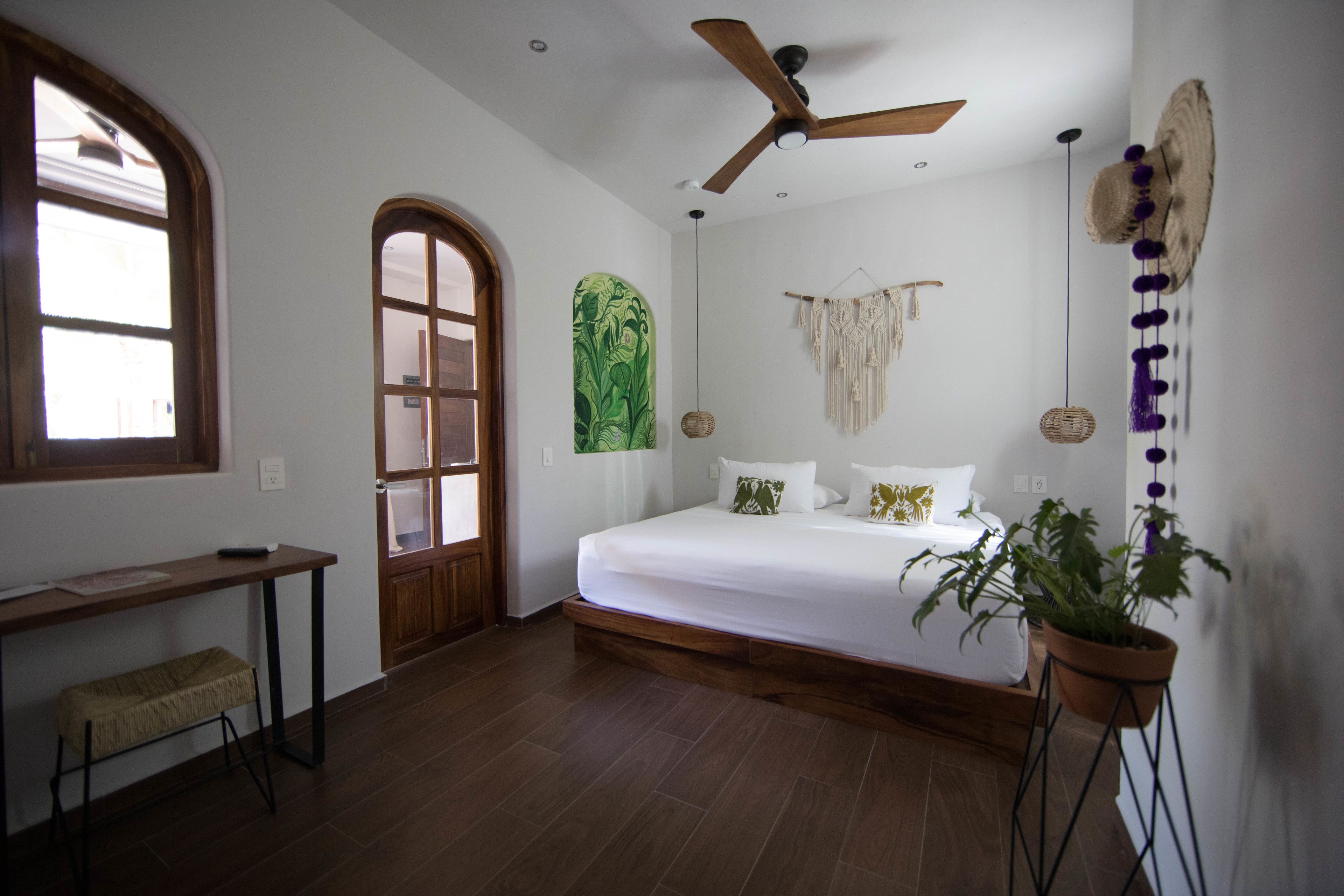 A bedroom with a bed, a ceiling fan, a large window, a door, a bench, and a potted plant.