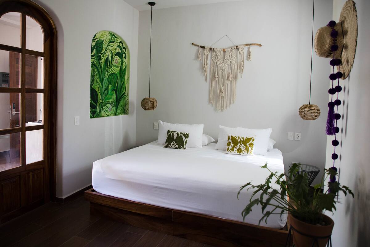 A bedroom with a large bed, a green abstract painting, a macrame wall hanging, and a potted plant.