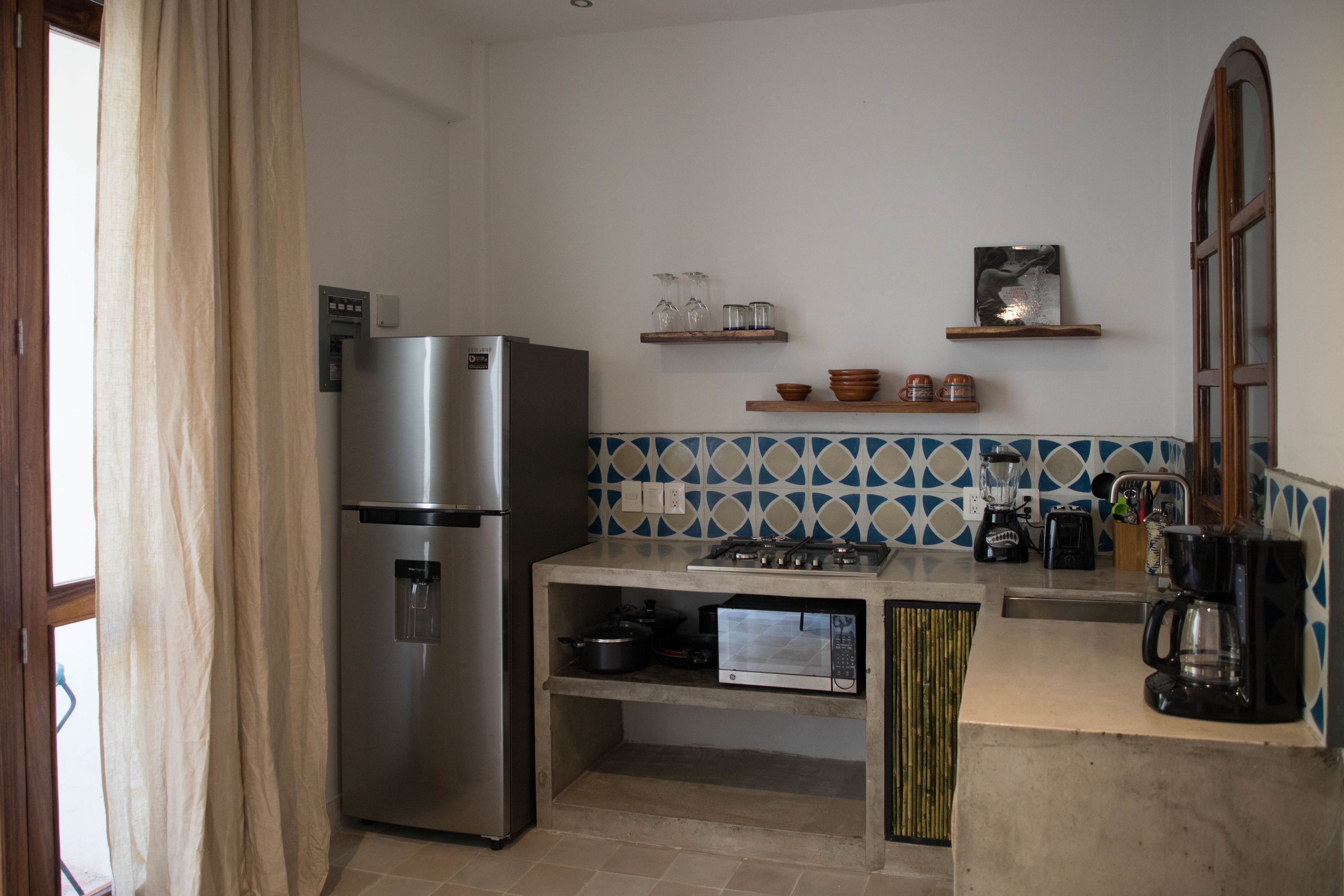 Cocina compacta con refrigerador, microondas, estufa a gas y cafetera. Se observan estantes con utensilios de cocina y una ventana con cortinas.