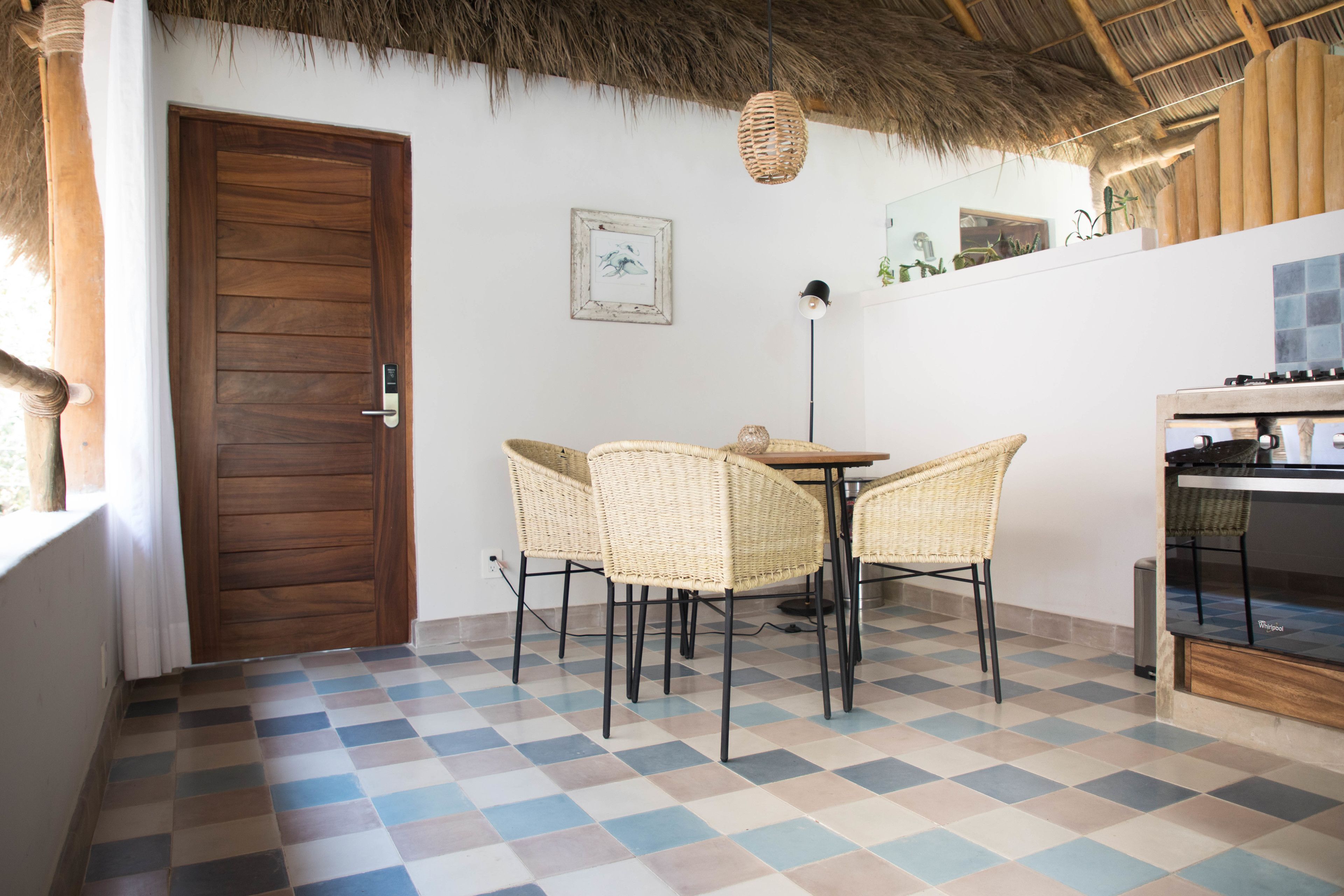 A room with a checkered floor, a wooden door, wicker chairs, a table, a hanging light, and a stove.