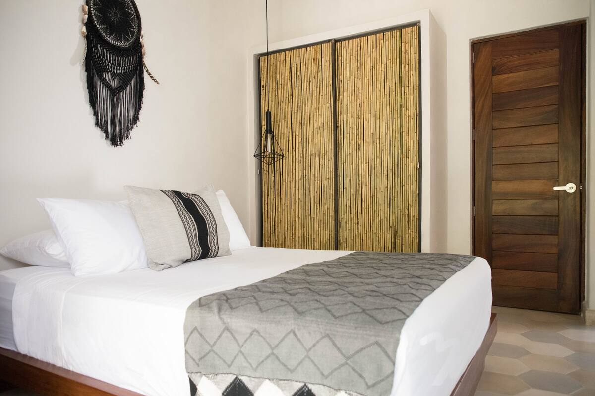 A neatly made bed with a patterned blanket, a wall-mounted macrame decoration, a bamboo-patterned wardrobe, and a wooden door.