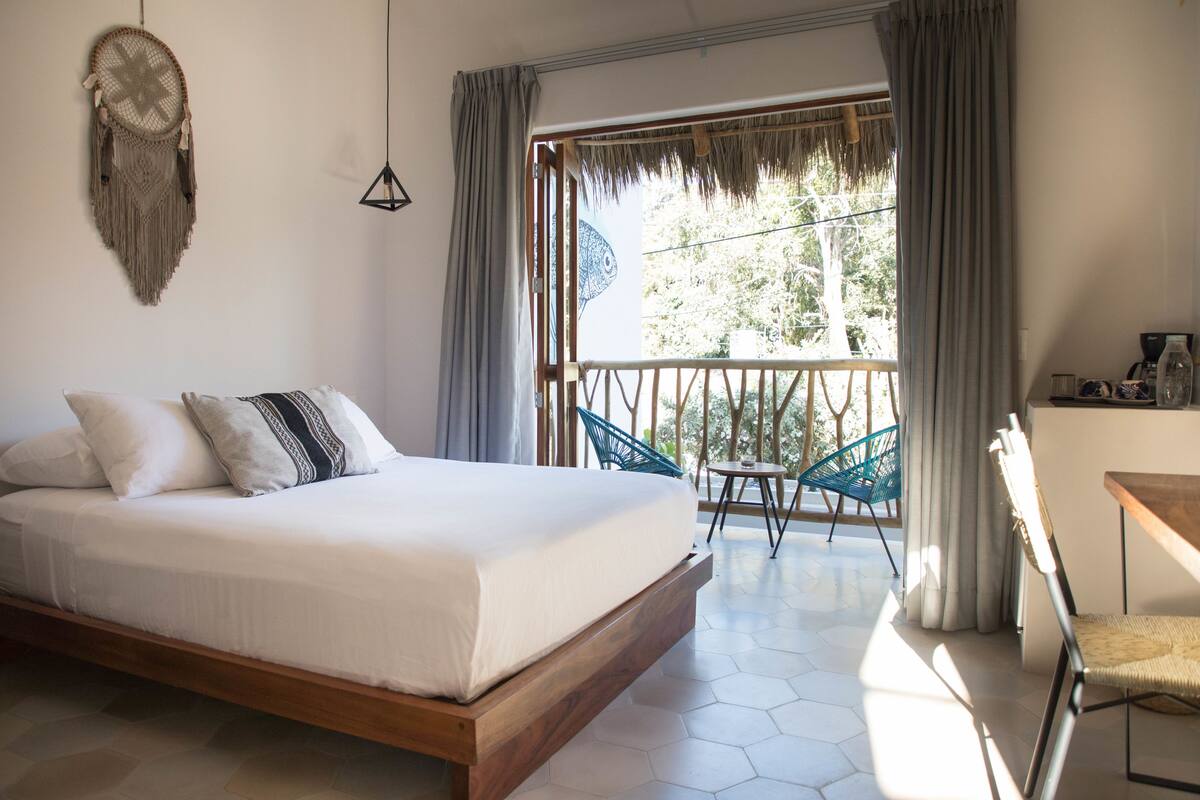 A bedroom with a bed, a balcony with chairs, and a view of trees.