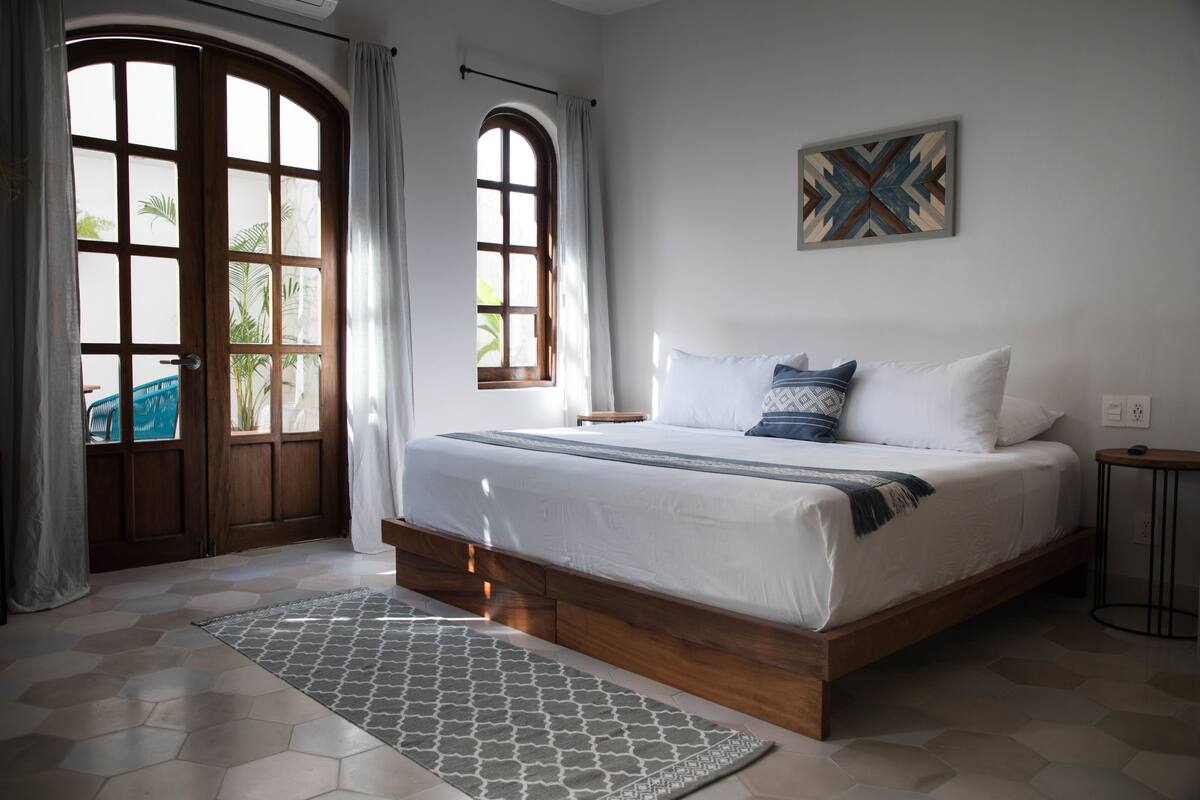 A bedroom with a wooden bed frame, a patterned rug, and a large window with curtains.