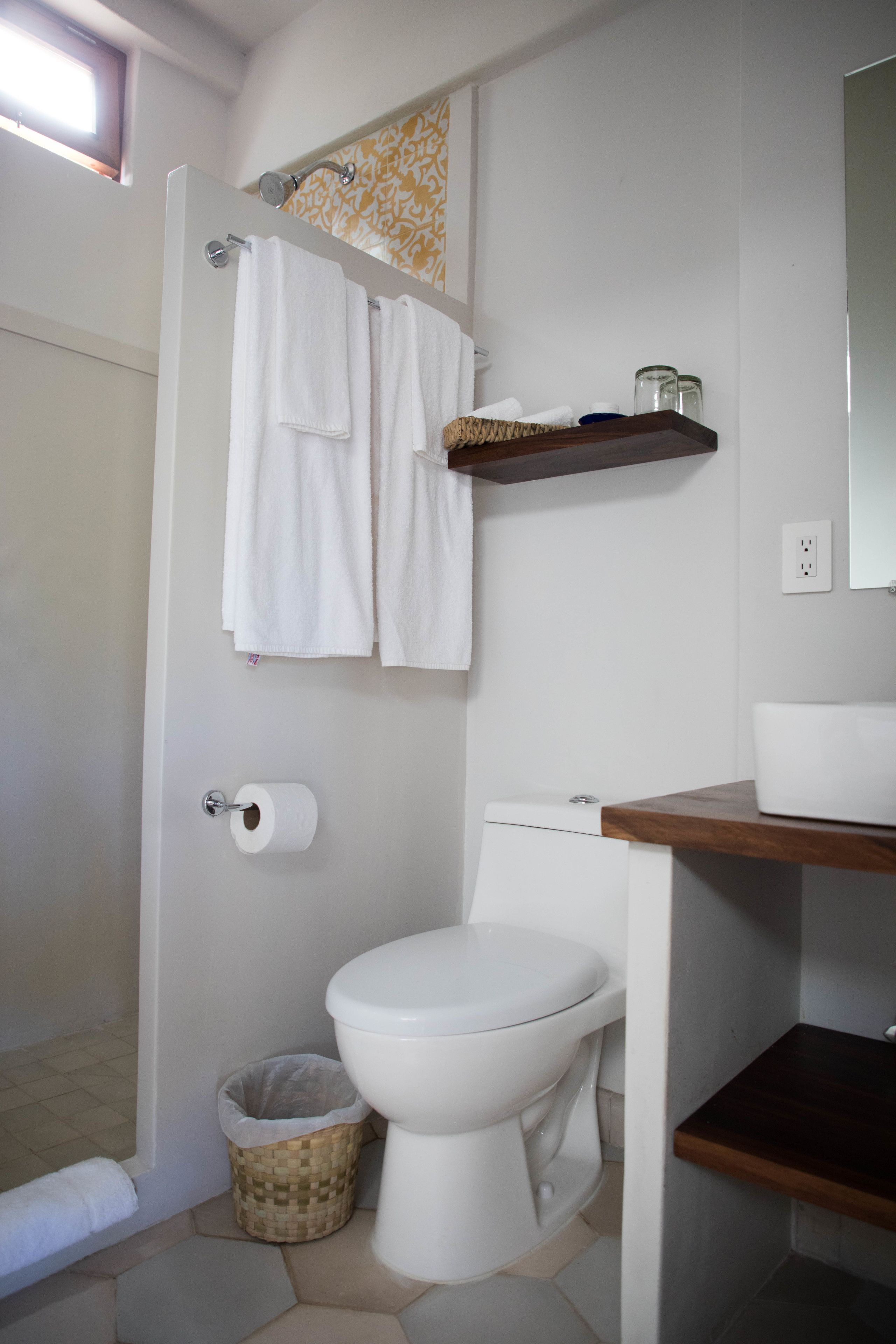 Un baño con inodoro, ducha, toallero con toallas, dispensador de papel higiénico, cesto de basura y estante de madera con espejo.