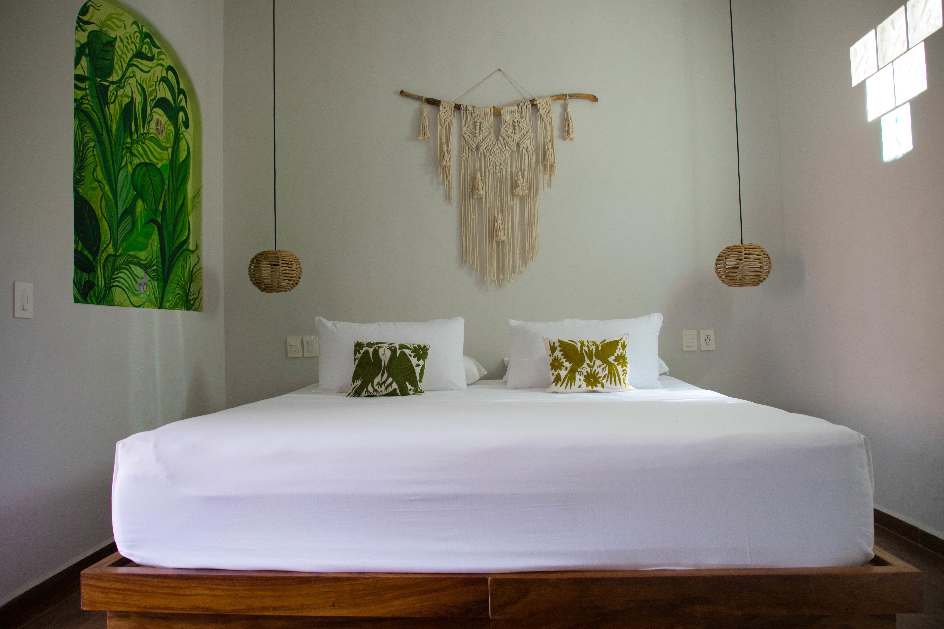 A bed with white bedding, two pillows with green and gold patterns, a macrame wall hanging, and a woven light fixture.