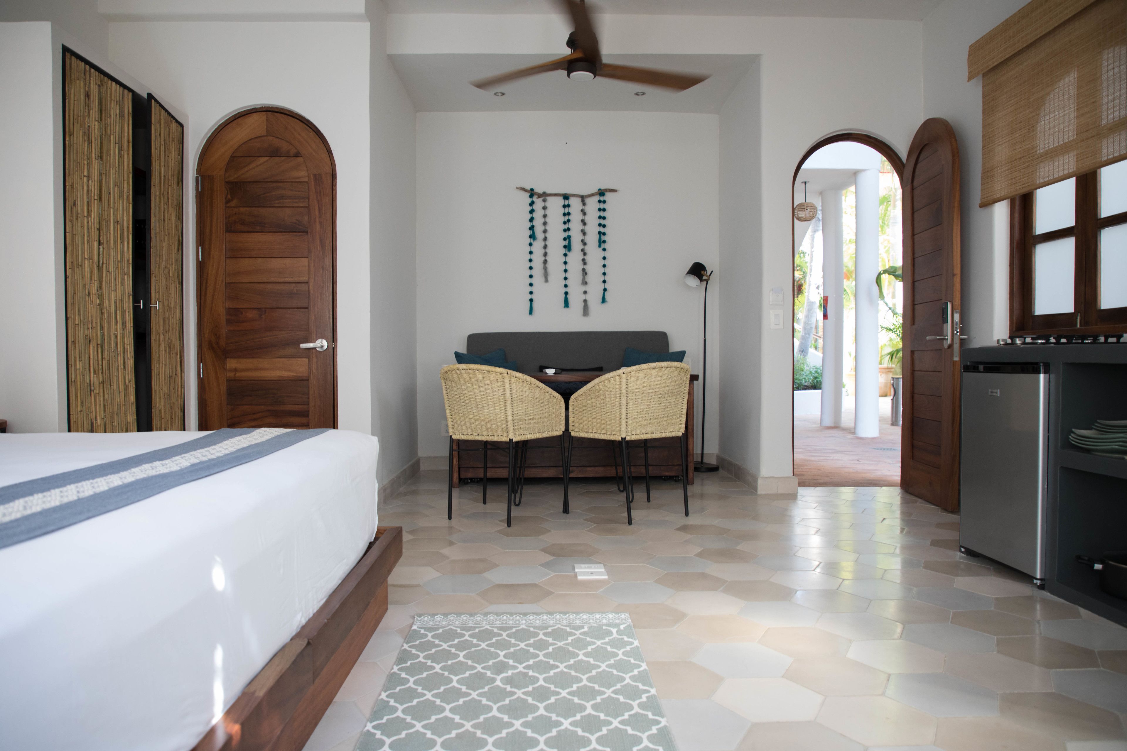 A hotel room with a bed, a dining area with two chairs, a ceiling fan, and a kitchen area.