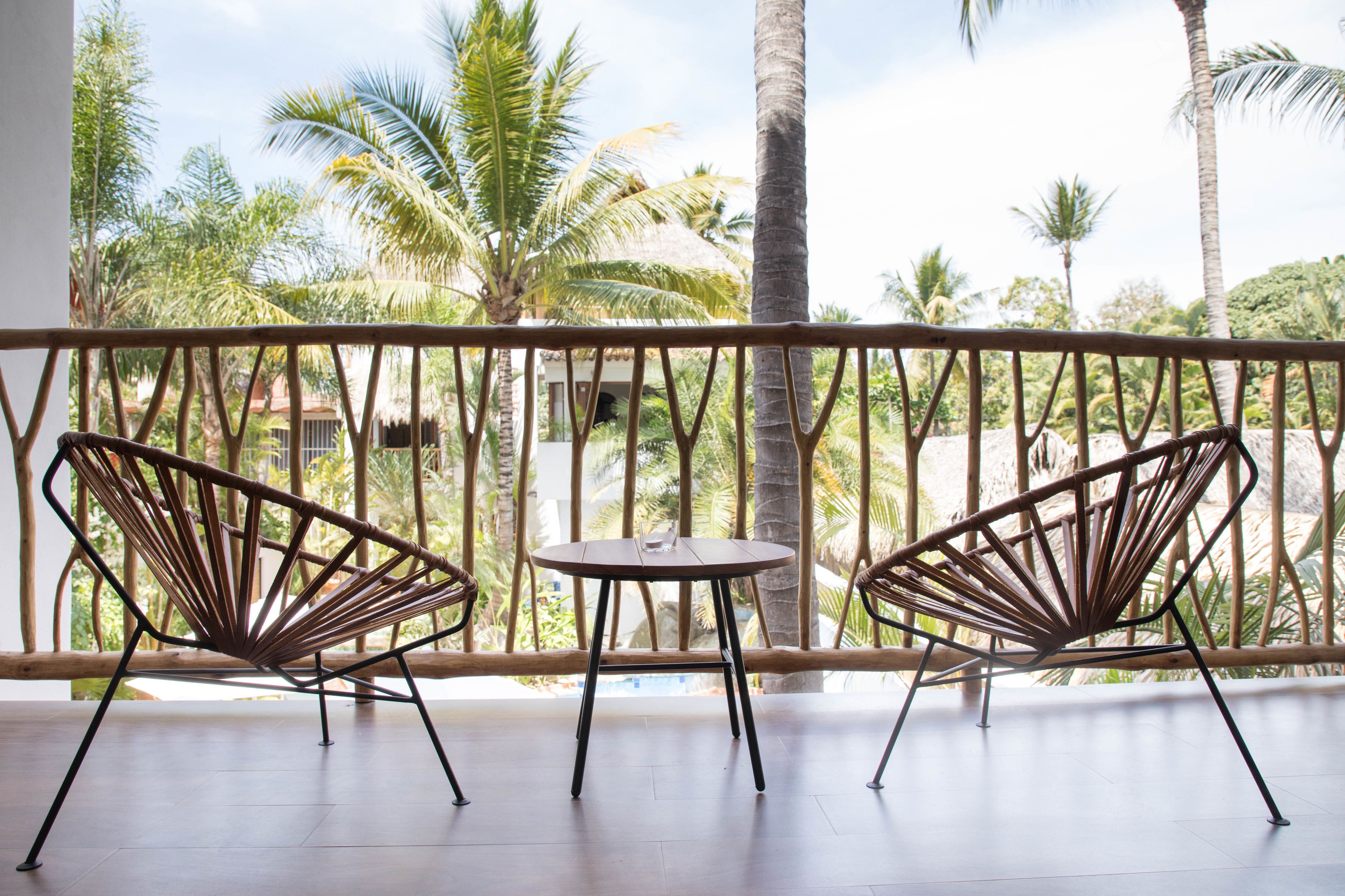 A balcony with two wicker chairs and a small round table, overlooking a tropical garden with palm trees.