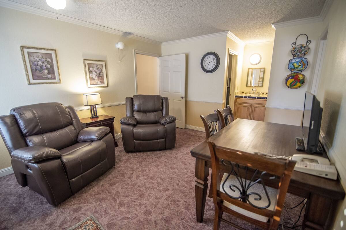 A living room with a brown leather sofa, a dining table with chairs, a TV, and a clock on the wall.