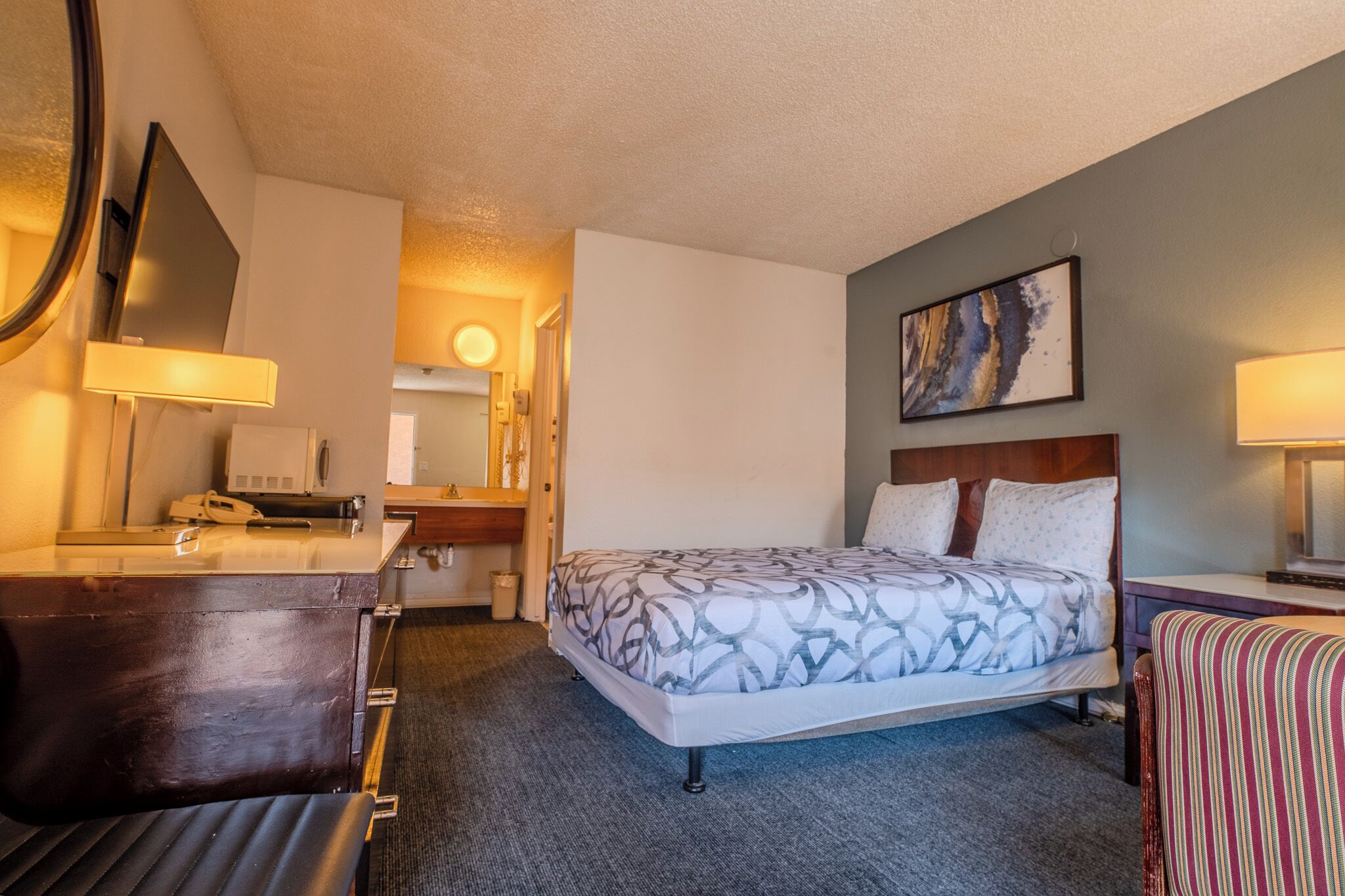 A hotel room with a bed, a desk, a chair, a mirror, and a painting on the wall.