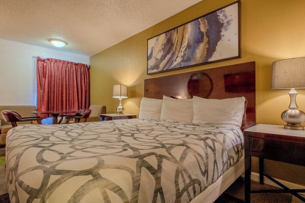 A hotel room with a large bed, a headboard, a bedside table with a lamp, a desk with a lamp, a chair, a small table, and a red curtain.