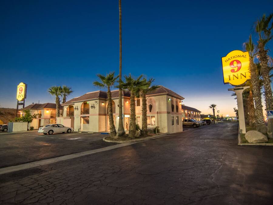 A hotel with a sign indicating it is a National 9 Inn, featuring a two-story building with a tiled roof, palm trees, and a paved driveway.