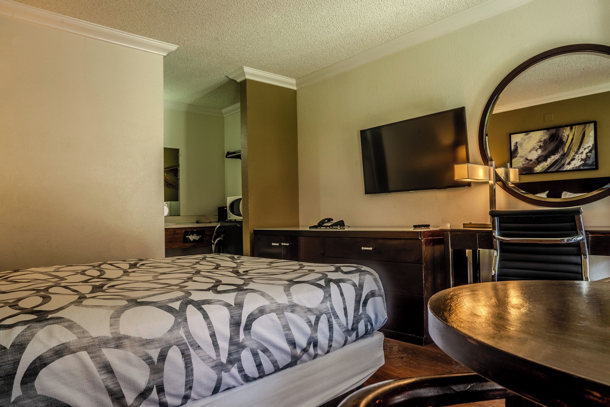 A hotel room with a large bed, a desk with a chair, a flat-screen TV, a round mirror, and a painting on the wall.