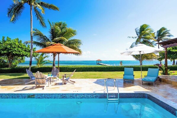 A poolside area with lounge chairs and umbrellas, a clear view of the ocean, and a well-maintained garden.