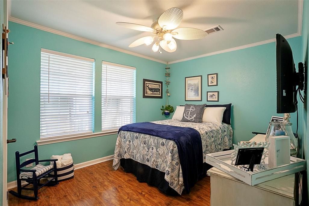 Une chambre à coucher comprenant un lit, un bureau, une chaise, un ventilateur de plafond et deux fenêtres.