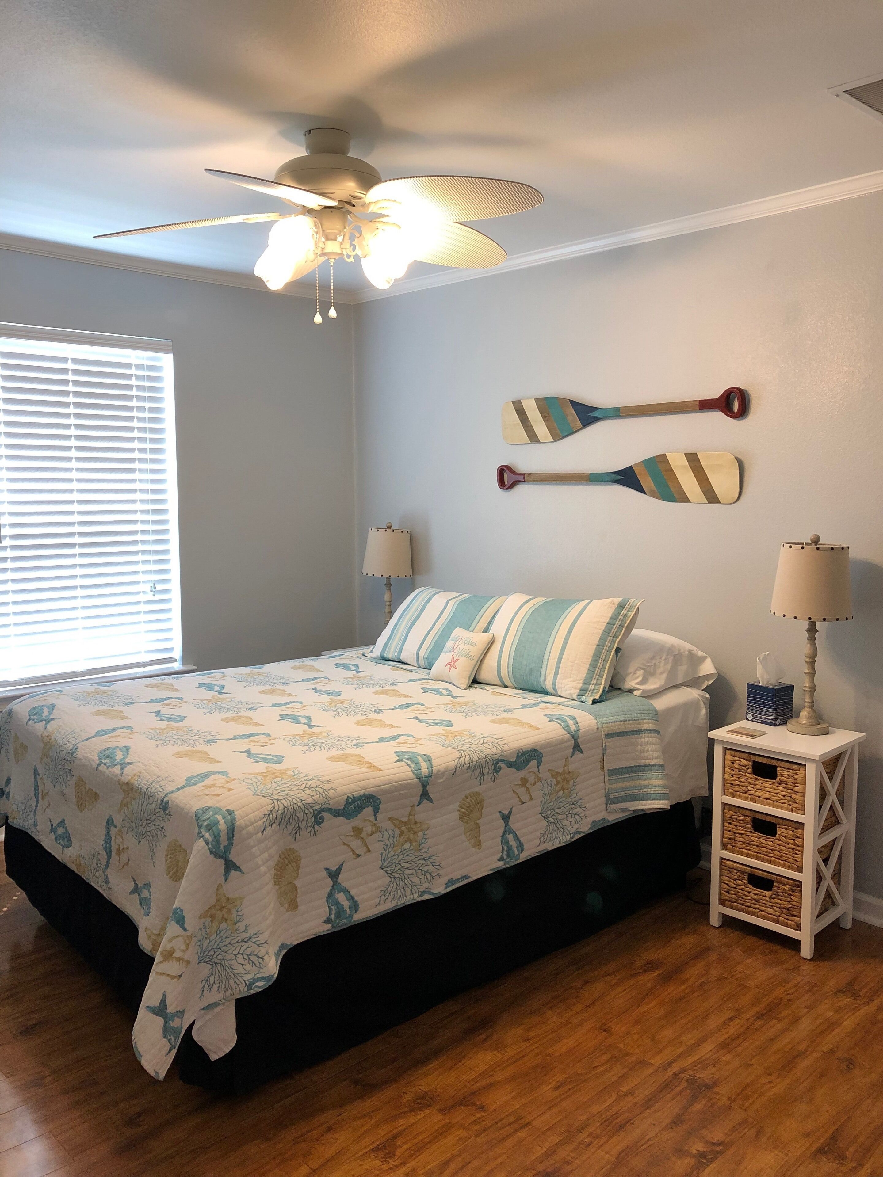 A bedroom with a bed, a ceiling fan, a window with blinds, and a nightstand with a lamp.