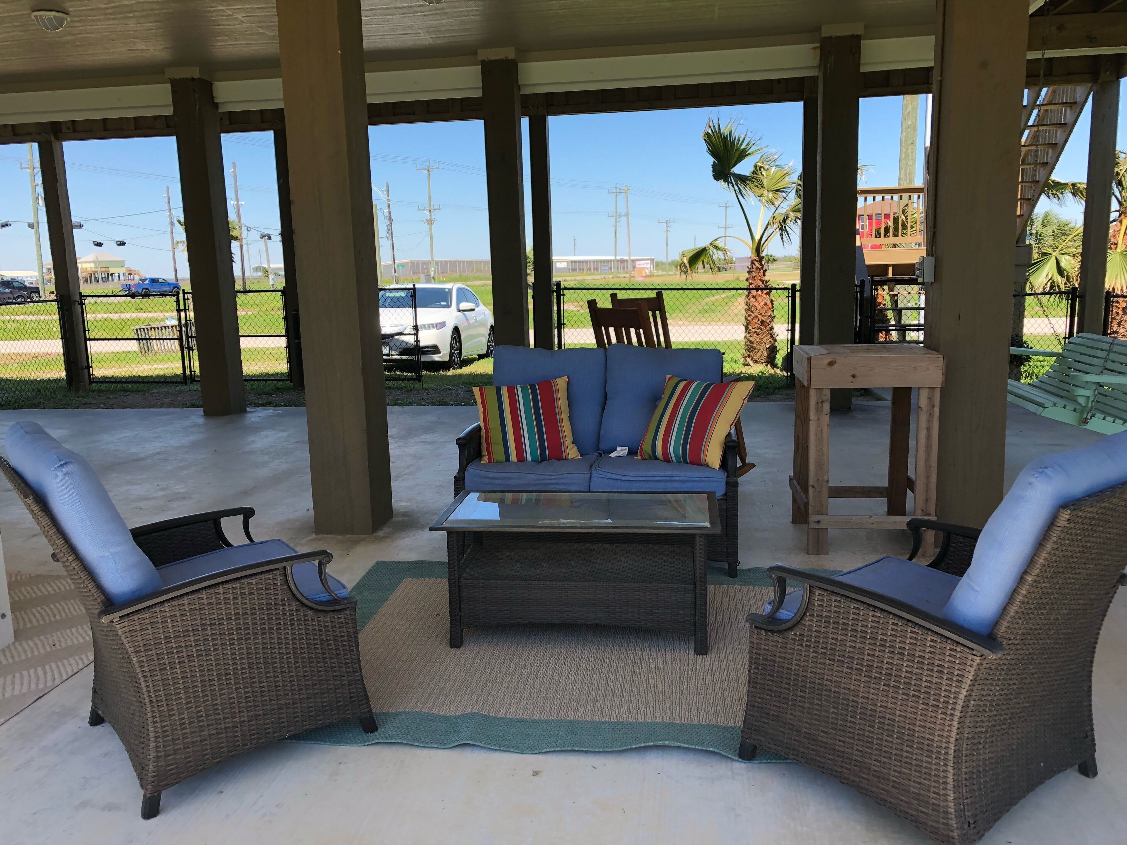 An outdoor covered seating area with wicker furniture, a glass-top coffee table, and a view of a parking lot and distant buildings.