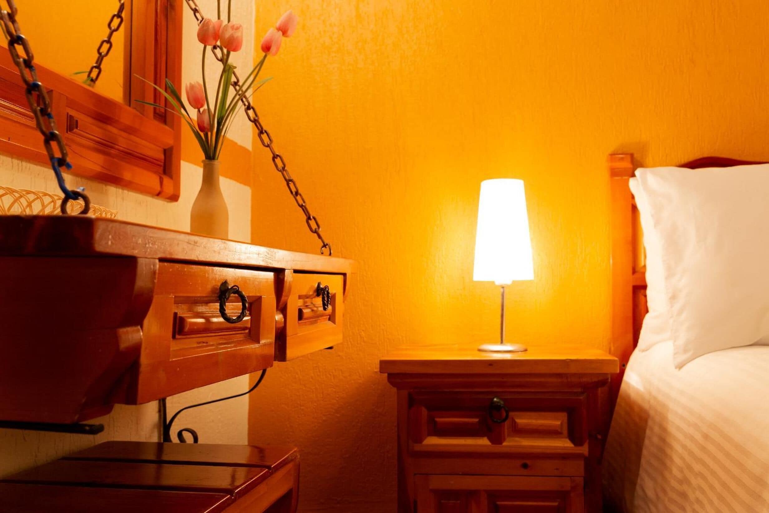 A hotel room with a wooden nightstand, a bedside table with a lamp, a bed with white bedding, and a vase with flowers on the nightstand.