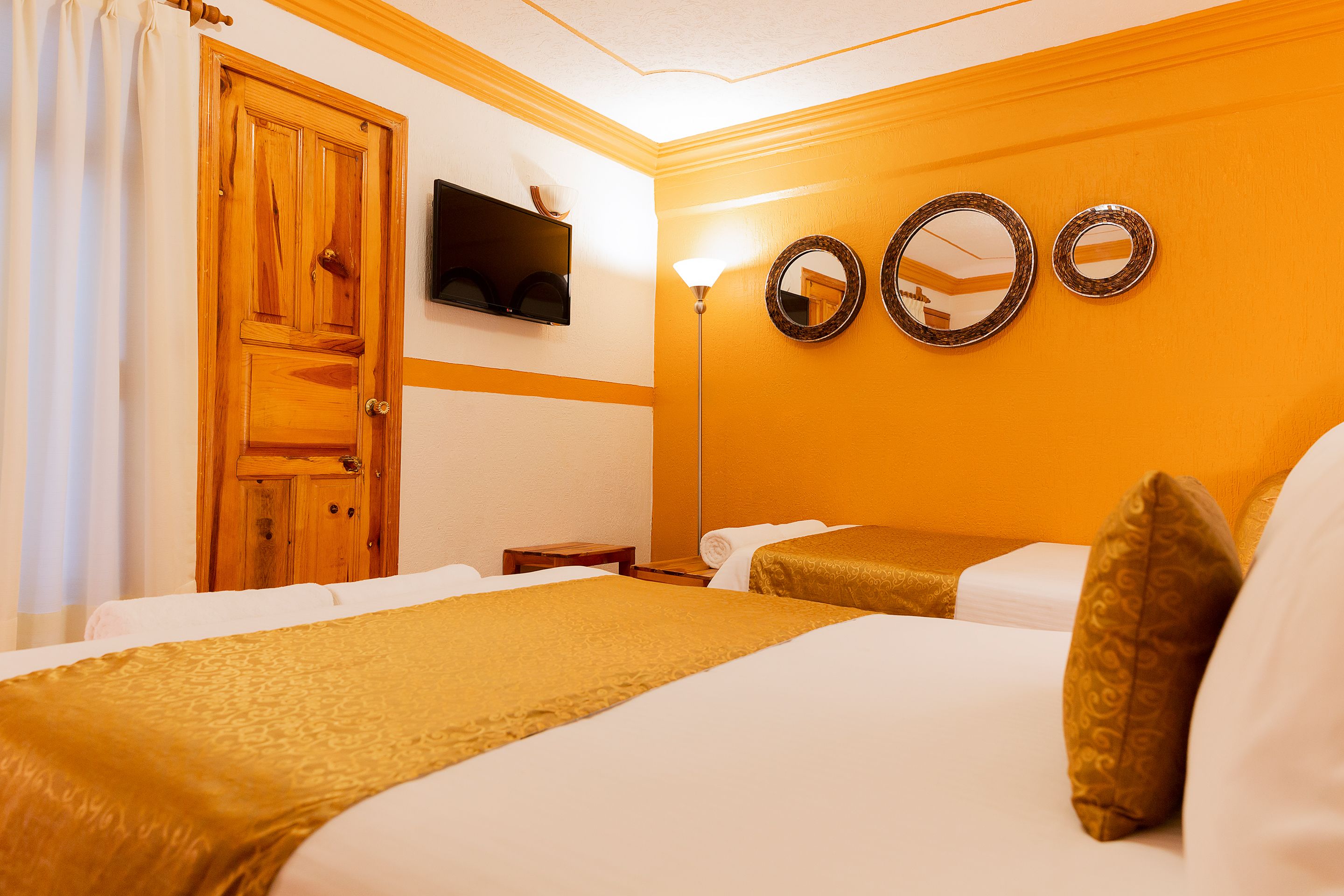 Habitación de hotel con dos camas, una puerta de madera, un televisor de pantalla plana y tres espejos adosados a la pared.
