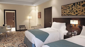 Quarto de hotel com duas camas, uma escrivaninha, uma cadeira, um abajur e uma obra de arte na parede.