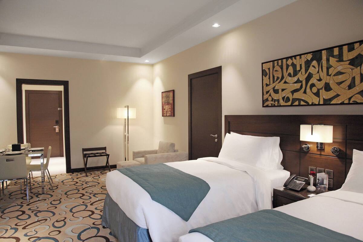 A hotel room with two beds, a desk, a chair, a lamp, and a wall art piece.