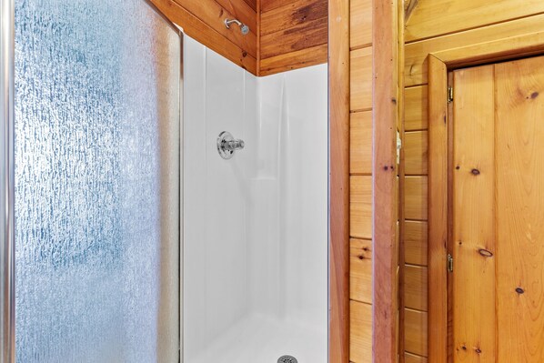Una puerta de madera con manija metálica, un box de ducha blanco con puerta de vidrio, y un techo de madera con rejilla de ventilación.