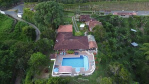 Oversigtsbillede af et resort med en stor swimmingpool, flere bygninger med røde tagsten og frodig vegetation omkring ejendommen.