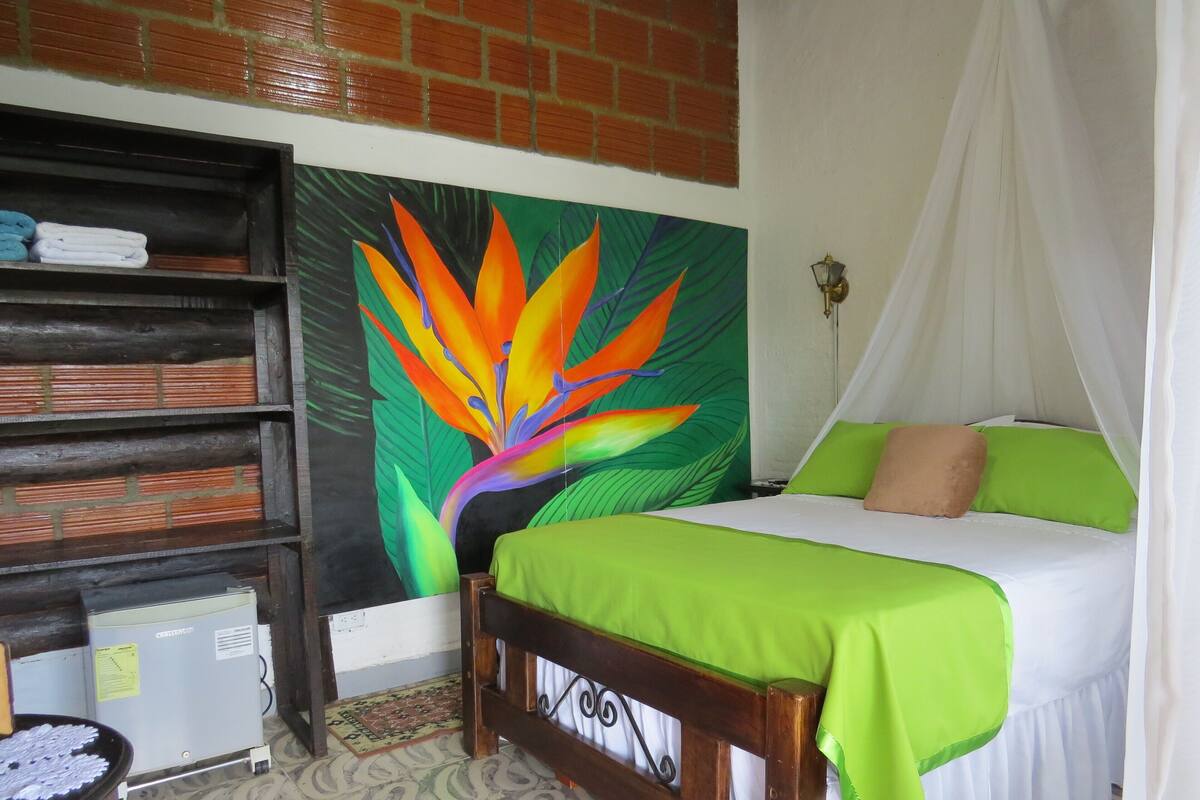 Ein Zimmer mit einem Bett, einem Regal mit gefalteten Handtüchern, einem Heizkörper und einem farbenfrohen Wandbild.