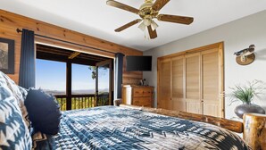 Un dormitorio con una cama grande, ventilador de techo, televisor y vistas al exterior.