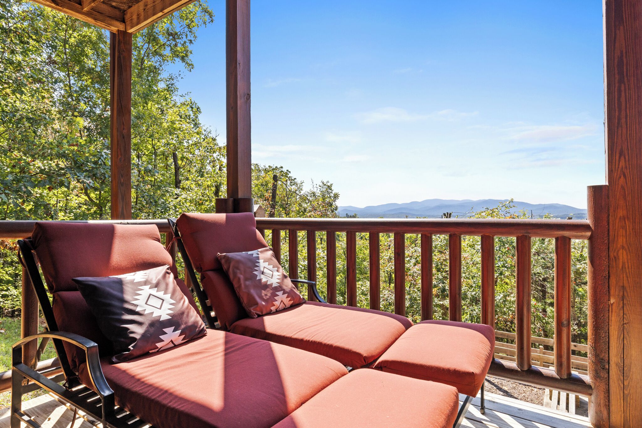 Una terraza de madera con dos sillas acolchadas y barandilla de madera, con vistas a un bosque y montañas distantes.
