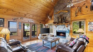 Interior acogedor de una cabaña de troncos con chimenea de piedra, sofás de cuero y una televisión.