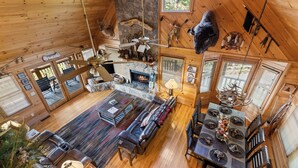 Un interior de cabaña espacioso con chimenea, pisos de madera, un sofá de cuero, una mesa de comedor y un ventilador de techo.
