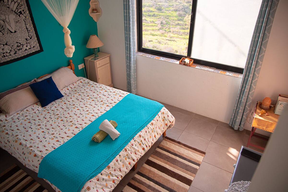 A bedroom with a bed, a nightstand, a lamp, a window with a view, and a small table with a book and a candle.