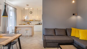 Ein modernes Wohnzimmer mit grauem Sofa, gelben Kissen, einem Holzessttisch und einer Küche im Hintergrund.
