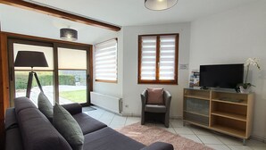 A living room with a dark grey sofa, a small armchair, a television on a wooden stand, and a large window with blinds.