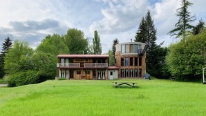 Un edificio de dos pisos con un balcón y una pequeña estructura adosada, rodeado de un campo cubierto de hierba y árboles.
