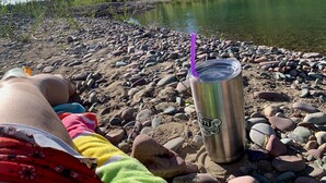 Una persona relajándose en una orilla rocosa con un vaso de acero inoxidable y una pajita morada.