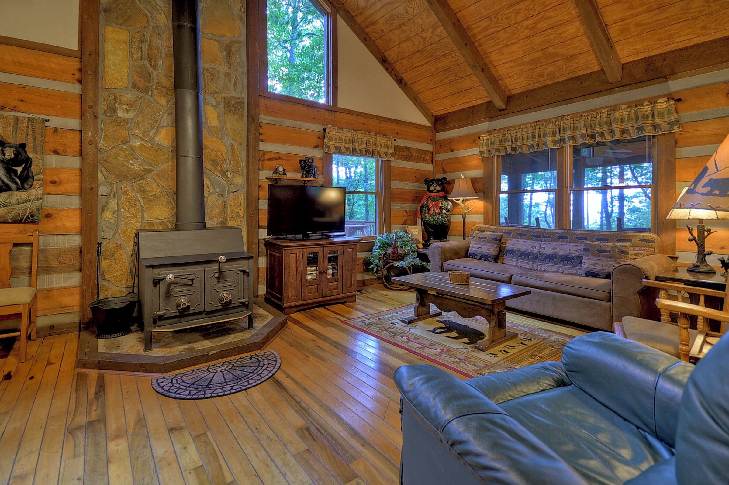A cozy cabin interior with a stone fireplace, wooden furniture, and large windows.