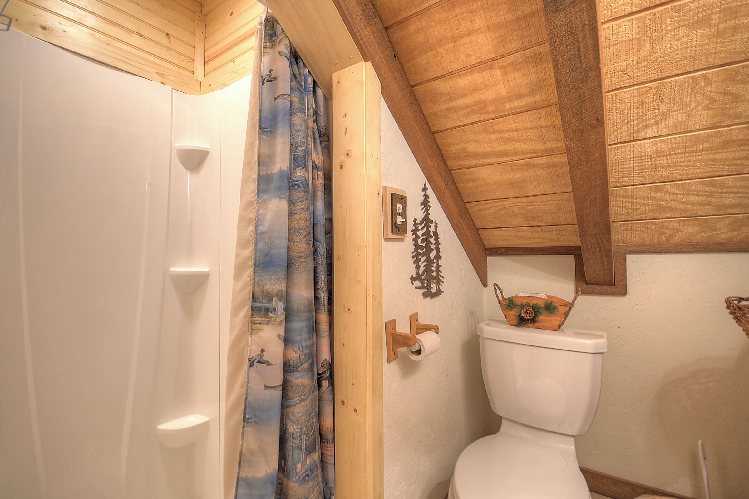 Une salle de bain avec un plafond en bois, des toilettes blanches et un rideau de douche orné d’un motif représentant une forêt.